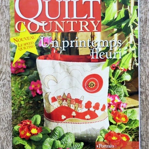 Peut inclure: La couverture du magazine "Quilt Country" présente un sac matelassé avec un motif rouge et blanc, sur fond de fleurs et de verdure. Le titre du magazine est en grandes lettres blanches sur fond rouge, avec du texte français en dessous.