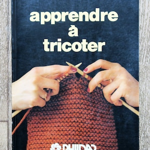 Peut inclure: Couverture de livre vintage avec le titre "apprendre à tricoter" en lettres blanches sur fond bleu foncé. L'image montre des mains tricotant avec des aiguilles en bois et de la laine orange. Le logo Phildar est en bas.