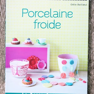 Könnte beinhalten: Ein Buchcover mit gelbem Hintergrund und einem türkisfarbenen Titel, der "Atelier Decouverte Porcelaine Froide" lautet, darunter der Name des Autors "Odile Bailloul". Das Cover zeigt ein rosa Regal mit einer weißen Kerze, einer weißen Tasse und einem kleinen Keramikkuchen mit roten Kirschen darauf. Eine Schnur aus bunten Keramikknöpfen hängt an der Tasse.