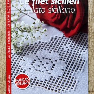 Peut inclure: Couverture de livre avec le titre "Le filet sicilien Sfilato siciliano" en blanc sur fond rouge. La couverture présente un motif de dentelle blanche, une rose rouge et des fleurs blanches. Le texte "FRANÇAIS ITALIANO" est dans un cercle rouge et blanc.