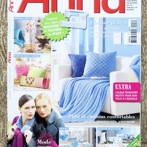 May include: A magazine cover featuring a blue knitted throw blanket and pillows on a white couch. The magazine is titled "Anna" and features the text "Plaid and cushions comfortable for cozy moments".