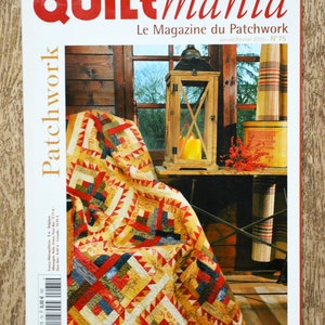 Pode incluir: Capa da revista "Quiltmania", com o subtítulo "Le Magazine du Patchwork". Apresenta um edredom colorido sobre uma cadeira, uma lanterna de madeira e uma janela. A revista está sobre uma superfície de madeira. Edição de janeiro/fevereiro de 2010.