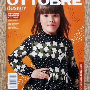 May include: Magazine cover with the title "OTTOBRE design" and a young girl wearing a black dress with white and olive green floral patterns. The magazine includes the text "kids fashion 62-170 cm" and the word "DEUTSCH". The background is orange.