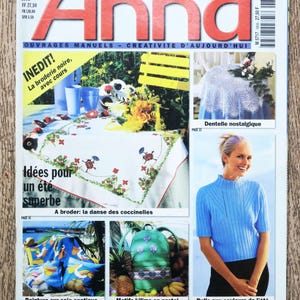 May include: A magazine cover for Anna, a French publication about crafting and home decor. The cover features a colorful illustration of ladybugs on a white tablecloth, a blue knitted sweater, and a green and white patterned backpack. The text on the cover reads "Anna, Une Revue Aenne Burda, Ouvrages Manuels - Creativite d'Aujourd'hui, Inedit! La broderie noire, avec cours, Idees pour un ete superbe, A broder: la danse des coccinelles, Dentelle nostalgique, Peinture sur soie exotique, Motifs kilims en pastel, Pulls aux couleurs de l'ete, Avec motif multi-decalquable et planche a ouvrages."