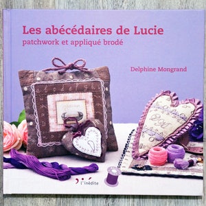 以下が含まれることがあります： 「Les abécédaires de Lucie」というタイトルの本で、副題は「patchwork et appliqué brodé」です。表紙は紫で、ハート型の枕や正方形の枕など、手作りの布工芸品の画像が掲載されています。
