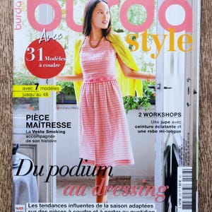 Op de afbeelding: Een tijdschriftcover met een vrouw die een roze-wit gestreepte jurk en een gele cardigan draagt. Het tijdschrift heet "burda style" en heeft de tekst "31 Modellen om te naaien" en "2 Workshops" op de cover.