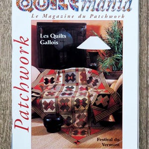 Op de afbeelding: De cover van het tijdschrift "Quiltmania" toont een patchworkquilt met rode, bruine en crèmekleurige geometrische patronen. De titel van het tijdschrift staat in kleurrijke letters, met de woorden "Patchwork" en "Les Quilts Gallois" zichtbaar.