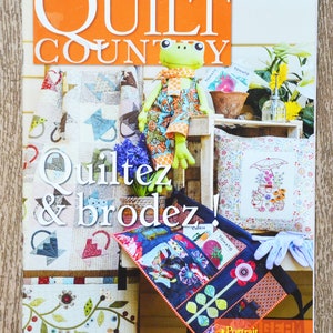 以下が含まれることがあります： 「Quilt Country」誌の表紙には、カエルの人形、キルト、そして「Quiltez & brodez！」という文字が掲載されています。雑誌には、白いタイトルが付いたオレンジ色のバナーがあります。その他のアイテムには、傘のデザインの枕とトートバッグが含まれています。