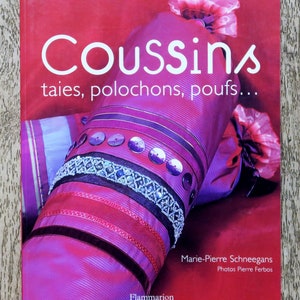 May include: A book titled "Coussins: taies, polochons, poufs..." by Marie-Pierre Schneegans with photographs by Pierre Ferbos. The book cover features a close-up of a purple and red cushion with decorative trim.