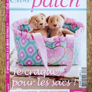 Op de afbeelding: Een magazine cover met een tote bag met een hartontwerp en twee teddyberen. De tas heeft een roze handvat en een blauw en roze patroon. De tekst "Je craque pour les sacs!" is te zien.
