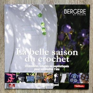 May include: Book cover featuring a white shirt with green crocheted buttons, a title that reads "La belle saison du crochet", and the text "20 modèles simples ou sophistiqués pour ensoleiller l'été".