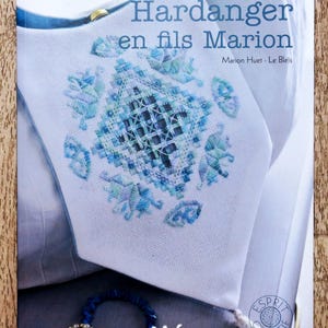 Pode incluir: Capa de livro com o título "Hardanger en fils Marion" de Marion Huet - Le Bleis. A capa apresenta um tecido branco com bordados azuis e um close-up de um colar de contas azuis e brancas.