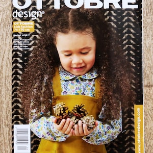 May include: A magazine cover featuring the title "OTTOBRE design" and a photograph of a young girl holding pine cones. The girl is wearing a floral shirt and a mustard-coloured overall dress. The magazine is labelled "kids fashion 74-170 cm".