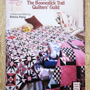 Op de afbeelding: De boekomslag toont "Quilts from The Booneslick Trail Quilters' Guild" door Bettina Havig. Het toont een quilt met geometrische patronen in roze, zwart, wit en rood. Andere quilts zijn op een tafel gestapeld, met een kussen en een hoed op het bed.