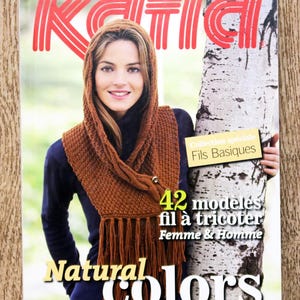 May include: A magazine cover featuring a woman wearing a brown knitted scarf and hood. The cover has the word "Katia" in red, and the text "Natural Colors" in yellow. The magazine is about knitting patterns.