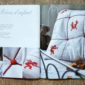 May include: A white pillow with red embroidered angels and red buttons. The pillow is on a white bed with a teddy bear and wooden blocks.