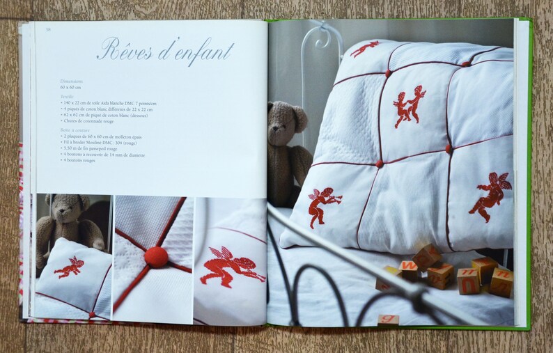 May include: A white pillow with red embroidered angels and red buttons. The pillow is on a white bed with a teddy bear and wooden blocks.