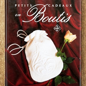 Può includere: Copertina del libro "Petits Cadeaux en Boutis" con un sacchetto bianco con coulisse e una rosa. Lo sfondo è un tessuto rosso scuro. Il nome dell'autore, Solange Kergreis, è in alto.