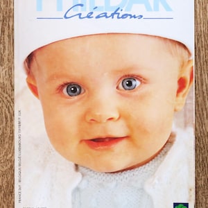 Könnte beinhalten: Ein Zeitschriften-Cover mit einem Baby mit blauen Augen, das einen weißen Hut und einen hellblauen Pullover trägt. Das Cover zeigt den Text "PHILDAR Créations" in Blau und die Worte "Layette PRINTEMPS-ÉTÉ".