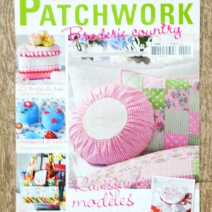 Op de afbeelding: Een tijdschriftcover met een roze en wit geruit kussen met een witte geborduurde cirkel in het midden. Het tijdschrift is getiteld "Elena Patchwork Broderie Country" en bevat de tekst "Ravissants modeles patchwork & broderie".