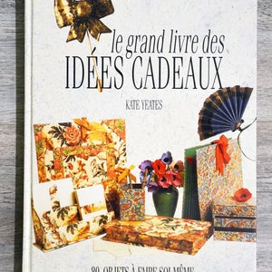 May include: A book titled "le grand livre des IDEES CADEAUX" by Kate Yeates. The book cover features a colorful illustration of various gift ideas, including a fan, a box, and a decorative paper holder. The text "80 OBJETS À FAIRE SOI-MÊME" is printed at the bottom of the cover.