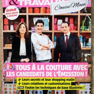 Può includere: Una copertina di rivista "MODES & TRAVAUX" in giallo e rosa. La copertina presenta tre persone davanti a scaffali pieni di forniture per cucire. Il testo include "Cousu Main" e "TOUS À LA COUTURE AVEC LES CANDIDATS DE L'ÉMISSION!"