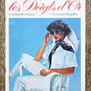 May include: A woman wearing a white shirt and white pants sits in a white wicker chair. She is wearing sunglasses and a blue scarf. The magazine cover reads "les Doigts d'Or encyclopédie pratique des travaux d'aiguilles" and "25 5FF - 30FB - 3.50FS".