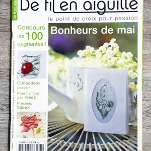 May include: A magazine cover for De fil en aiguille, a cross-stitch magazine. The cover features a white background with the title "De fil en aiguille" in black text. The subtitle is "le point de croix pour passion" in smaller black text. The main image is a white ceramic watering can with a white and green cross-stitch design of lily of the valley flowers. The text "Bonheurs de mai" is in large black text. The magazine cover also features a contest announcement for "les 100 gagnantes!" and a collection of cross-stitch designs.