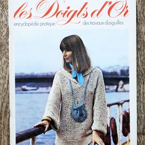 Peut inclure: Une couverture de magazine représentant une femme portant un pull marron clair avec un sac en crochet bleu. Le titre du magazine est "les Doigts d'Or" et le sous-titre est "encyclopédie pratique des travaux d'aiguilles".