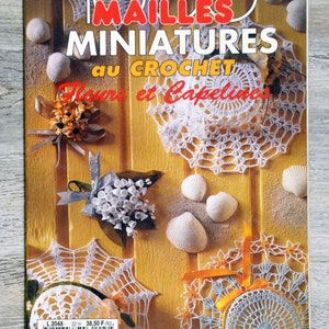 May include: A book cover with the title "1000 Mailles Miniatures au Crochet Fleurs et Capelines" in red and yellow text. The cover features a variety of crocheted flowers and doilies, along with seashells on a wooden background.
