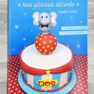 May include: A blue book with a white and red star pattern on the cover. The book title is "Mes gâteaux décorés" with the subtitle "Angélica Aublet". The cover features a cake decorated with a red, white, and blue circus theme. The cake is topped with a red ball with yellow stars and a gray elephant figurine.