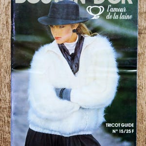 May include: A vintage knitting pattern booklet titled BOUTON D'OR l'amour de la laine TRICOT GUIDE N° 15/25 F. The cover features a person wearing a fuzzy white long-sleeve sweater, a dark gray scarf, and a dark blue fedora-style hat.