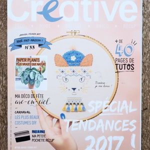 Peut inclure: Une couverture de magazine avec le titre "Creative" en grandes lettres blanches. La couverture présente un projet de broderie coloré d'un chat avec un chapeau bleu et un collier à glands jaunes et bleus. Le magazine s'intitule "Creative" et le texte de la couverture dit "100% Fait-Maison!" et "Special Tendances 2017!"