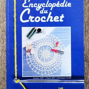 Peut inclure: Un livre bleu avec le titre "Encyclopédie du Crochet" et le sous-titre "Suzanne NEIL, Claude REN, Dominique CLAM, LTA LE TEMPS APPRIVOISÉ". La couverture du livre présente un napperon crocheté blanc, du rouge à lèvres et un crochet.