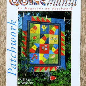 Op de afbeelding: De cover van het tijdschrift "Quiltmania" toont een kleurrijke patchworkquilt die buiten hangt. De quilt heeft een blauwe rand en een centraal ontwerp met bloemmotieven. De titel van het tijdschrift is in een veelkleurig, decoratief lettertype. De tekst "Le Magazine du Patchwork" is ook zichtbaar.