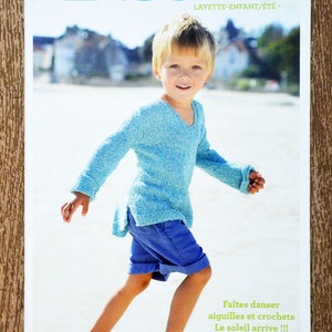May include: A magazine cover featuring a young boy wearing a light blue knitted sweater and blue shorts, standing on a beach. The magazine title is "PLASSARD LAYETTE-ENFANT/ÉTÉ - WISS" and the text "Faîtes danser aiguilles et crochets Le soleil arrive!!!" is at the bottom of the cover.