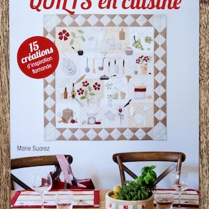 Peut inclure: Couverture de livre avec le titre "QUILTS en cuisine" en lettres rouges. La couverture présente une courtepointe colorée sur le thème de la cuisine, avec divers ustensiles de cuisine, de la nourriture et des fleurs. Le livre est de Marie Suarez et publié par les éditions de saxe.
