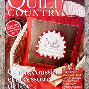 Pode incluir: Uma capa de revista vermelha e branca com o título "Quilt Country" e uma imagem de um travesseiro marrom e branco com um desenho de boneco de neve. O texto "Snowman Make me Smile" está no travesseiro. A capa da revista também apresenta o texto "Quilts, coussins et accessoires de fete" e uma lista de nomes sob o título "Portraits Exclusifs".