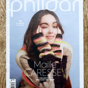 Könnte beinhalten: Ein Magazin-Cover mit einer Frau, die zwinkert und einen gestreiften Pullover und Armstulpen trägt. Das Magazin trägt den Titel "Phildar" mit dem Text "Maille CARESSE!" und "COLLECTION SPECIALE DOUCEUR". Der Hintergrund ist hellblau.