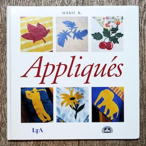 Peut inclure: Un livre intitulé "Appliqués" de Marie K. présentant des motifs d'appliqués colorés, dont une feuille rouge, une plante de tomate bleue, une branche de cerises, un golfeur, une fleur jaune et un éléphant. La couverture du livre présente également les logos LTA et DMC.