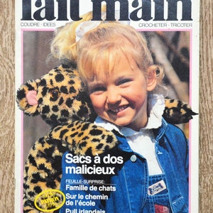 May include: A magazine cover featuring a young girl holding a leopard print stuffed animal. The magazine is titled "fait main" and has the words "Coudre - Idees" and "Crocheter - Tricoter" printed on the cover. The magazine cover also features the text "Sacs à dos malicieux" and "Feuille-Surprise: Famille de chats Sur le chemin de l'école Pull irlandais".
