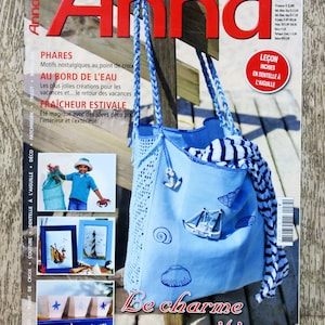 May include: A blue and white tote bag with a crocheted handle and a nautical theme. The bag is decorated with seashells, a sailboat, and an anchor. The text "Be charme maritime" is printed on the bag.