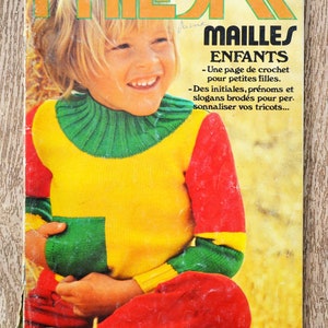 Pode incluir: Uma capa de revista com uma criança pequena vestindo um suéter de tricô colorido. A revista é intitulada "Phildar Mailles Enfants" e apresenta instruções para crochê e bordado de roupas infantis.