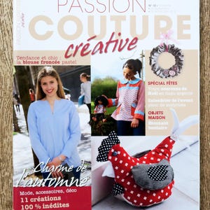 Peut inclure: Couverture du magazine "Passion Couture Créative" avec une femme en chemisier bleu. Présente un poulet en tissu, une couronne et le texte : "Charme de l'automne". Le magazine traite de mode, d'accessoires et de décoration intérieure.