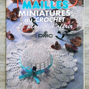 May include: A magazine cover featuring crochet projects. The title reads "100 Mailes Miniatures au Crochet". The cover showcases white crochet items, including a hat with a blue ribbon and purple roses, and a doily. Dried flowers are scattered around.
