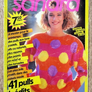 Op de afbeelding: Een tijdschriftcover met een vrouw die een kleurrijke trui met stippen draagt. Het tijdschrift heet "Sandra" en de tekst op de cover luidt "Les Plus Beaux Tricots" en "41 pulls inédits".