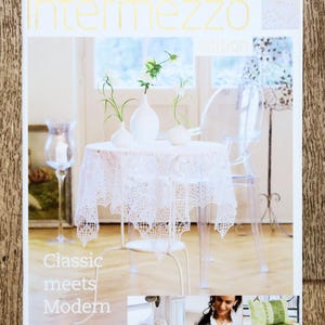 Può includere: Una copertina di rivista con il titolo "intermezzo edition" in lettere dorate. La copertina presenta un tavolino bianco con una tovaglia di pizzo, tre vasi bianchi con piante verdi e una sedia trasparente. È visibile anche il testo "Classic meets Modern".