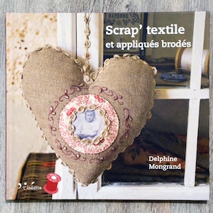 以下が含まれることがあります： デルフィーヌ・モングラン著「Scrap' textile et appliqués brodés」というタイトルの本。ハート型の麻のクッションに額縁に入った写真と刺繍が施されています。