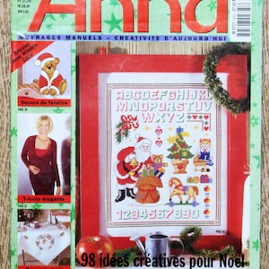 Op de afbeelding: Een tijdschriftcover met een rood en wit kruissteekontwerp van een kerstscène met de kerstman, een kerstboom en cadeaus. Het tijdschrift heet "Anna" en de tekst "98 idées créatives pour Noël" staat op de cover.