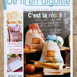 Peut inclure: Une couverture de magazine intitulée De fil en aiguille présentant deux sacs à cordon beiges avec des motifs au point de croix d'enfants tenant une grille. Les sacs sont posés sur une surface en bois avec des livres, une pomme et un tableau noir.